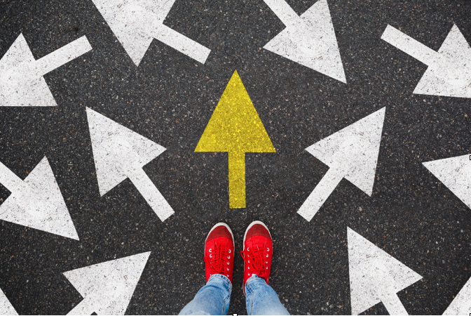 A view of jeans and red sneakers standing in front of several white arrows on asphalt pointing in different directions with a yellow arrow point forward.