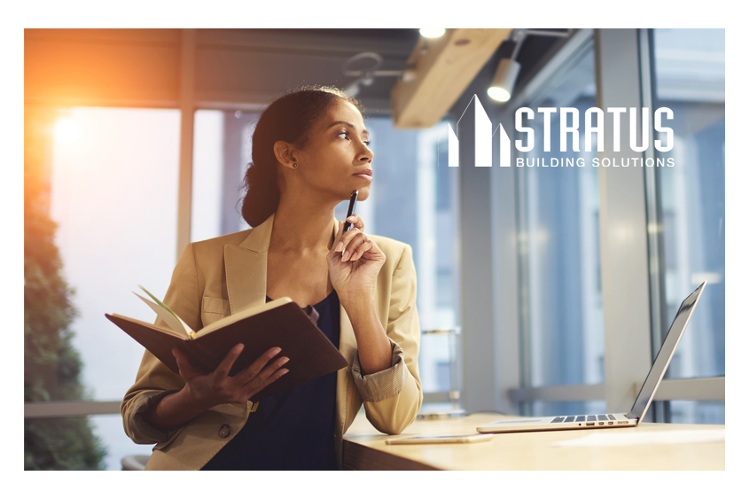 A woman in business attire stands next to a desk thoughtfully holding a pen up to her chin while holding a notebook