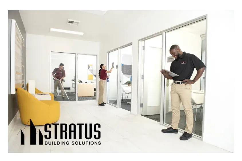 A Stratus Franchise Owner Smiles and Looks at a Tablet While Two Workers in the Background Vacuum and Wipe a Sliding Glass Door
