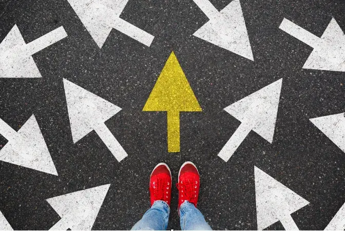 A view of jeans and red sneakers standing in front of several white arrows on asphalt pointing in different directions with a yellow arrow point forward.