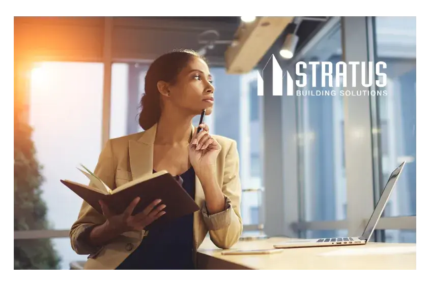 A woman in business attire stands next to a desk thoughtfully holding a pen up to her chin while holding a notebook